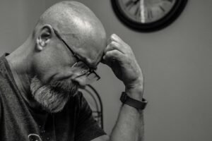 Black and white portrait of a thoughtful bald man indoors, capturing a moment of reflection with a wall clock in the background.