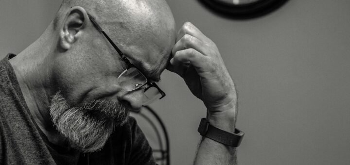 Black and white portrait of a thoughtful bald man indoors, capturing a moment of reflection with a wall clock in the background.