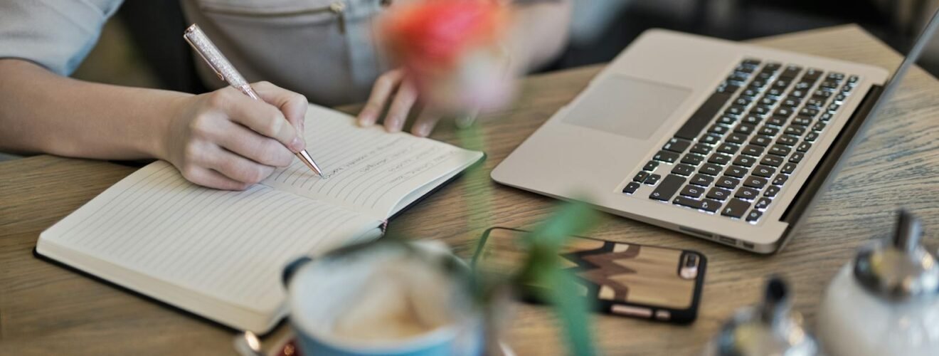 Woman writing in a notebook with a laptop and coffee cup on a desk. Ideal for workspace inspiration.