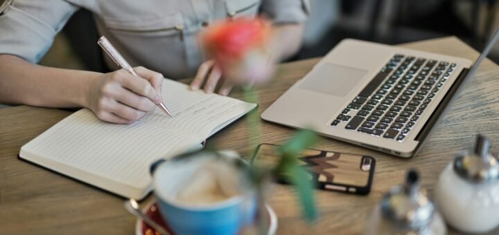 Woman writing in a notebook with a laptop and coffee cup on a desk. Ideal for workspace inspiration.