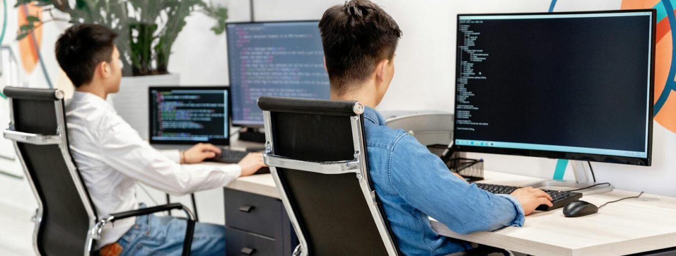 Two male developers at desks programming in a modern office workspace with large monitors.