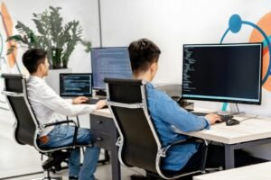 Two male developers at desks programming in a modern office workspace with large monitors.