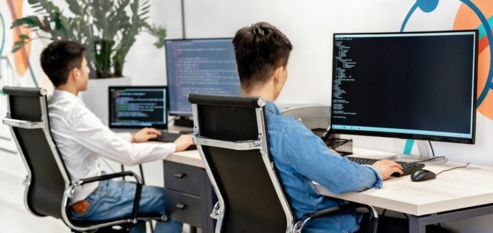 Two male developers at desks programming in a modern office workspace with large monitors.