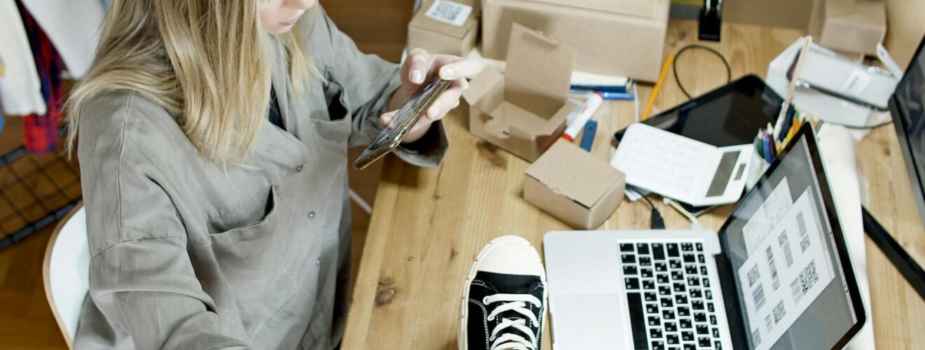 Woman photographing shoes for online sale in home workspace with laptop and packages.