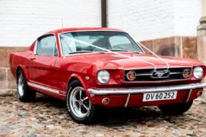 Vintage red Mustang with white ribbons parked on a cobblestone street.