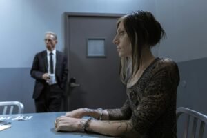 A handcuffed woman in an interrogation room with a detective in the background. Intense, dramatic scene.